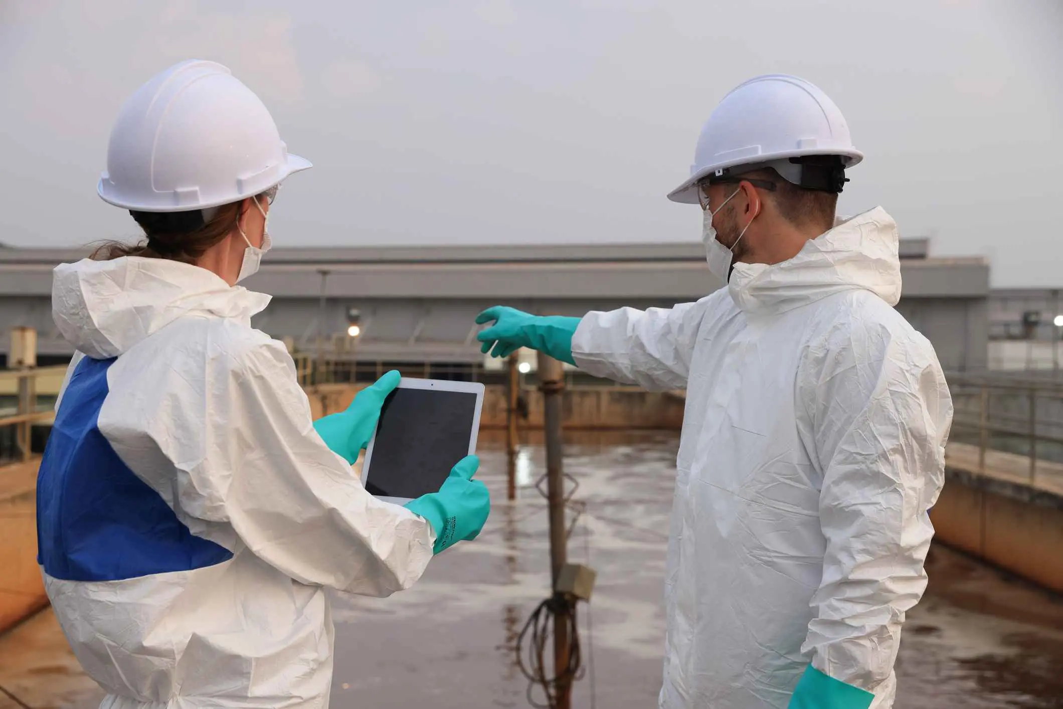 Two people in protective gear and helmets stand at a wastewater treatment facility; one holds a tablet while the other points.