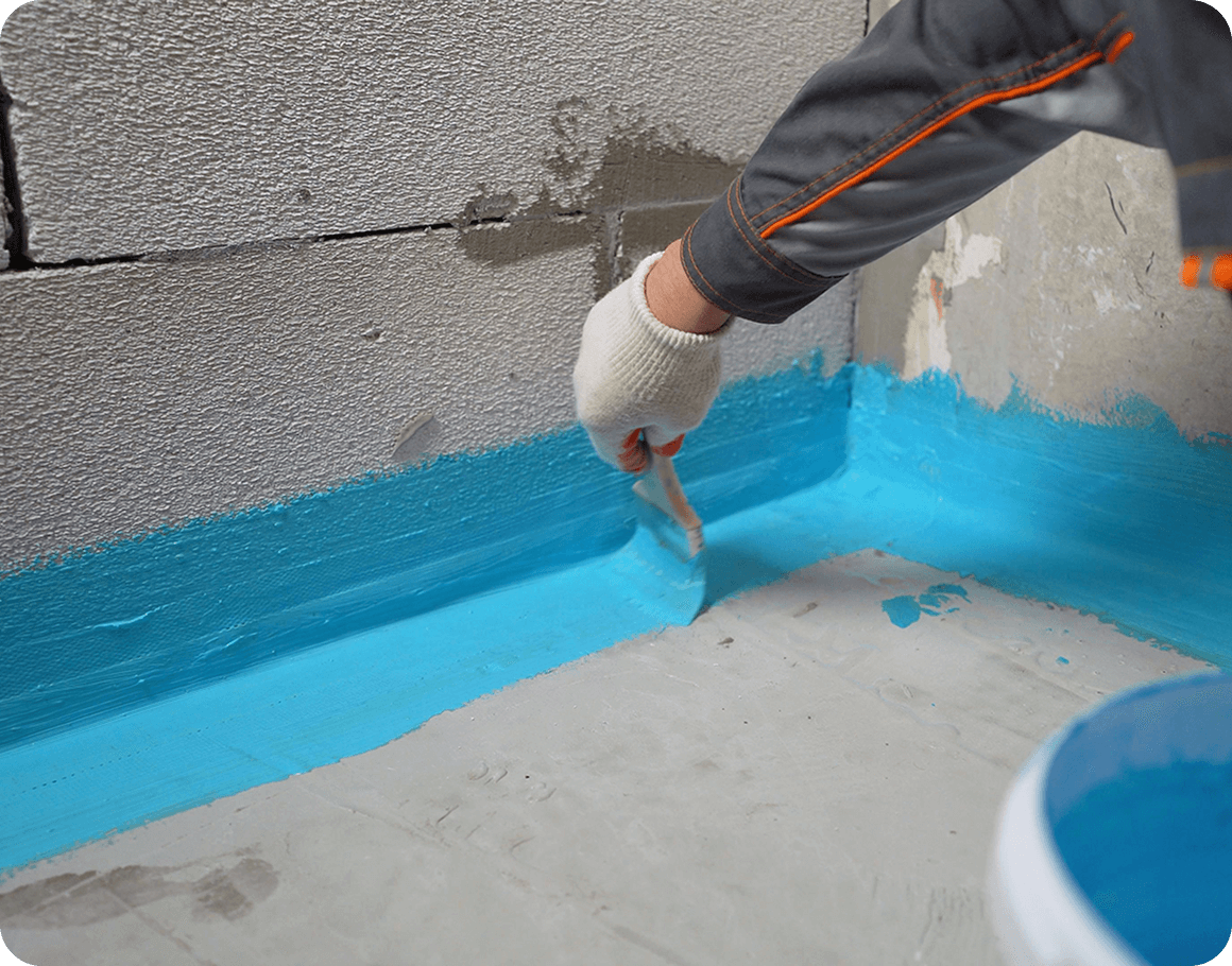 A person in gloves applies blue waterproofing paint to the corner of a concrete wall and floor.