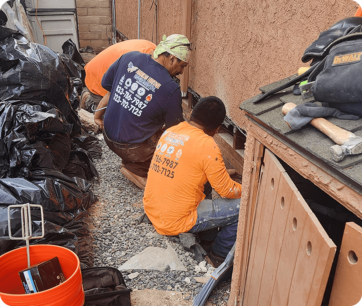 Two workers in orange shirts repairing a wall outdoors, surrounded by tools and black bags.