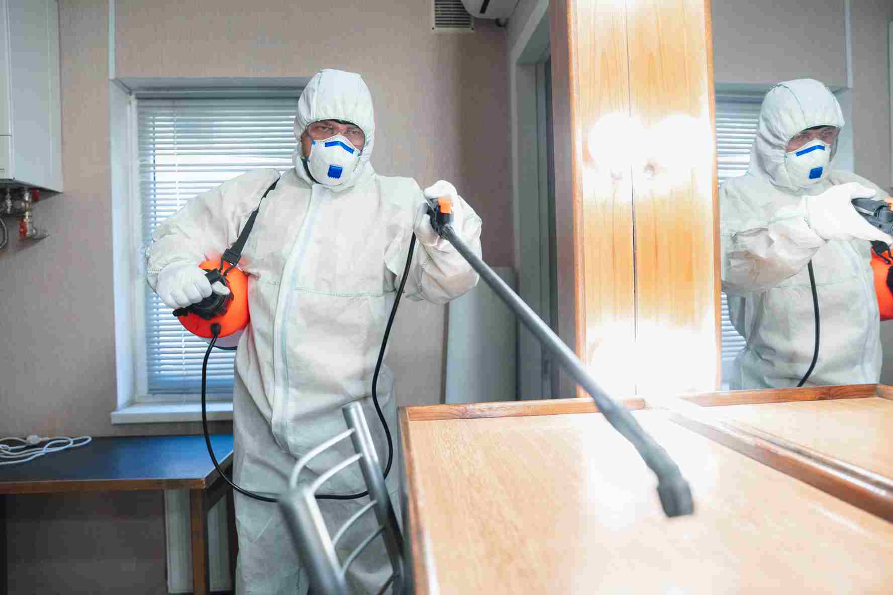 Person in protective suit and mask disinfecting a room with a spray device.