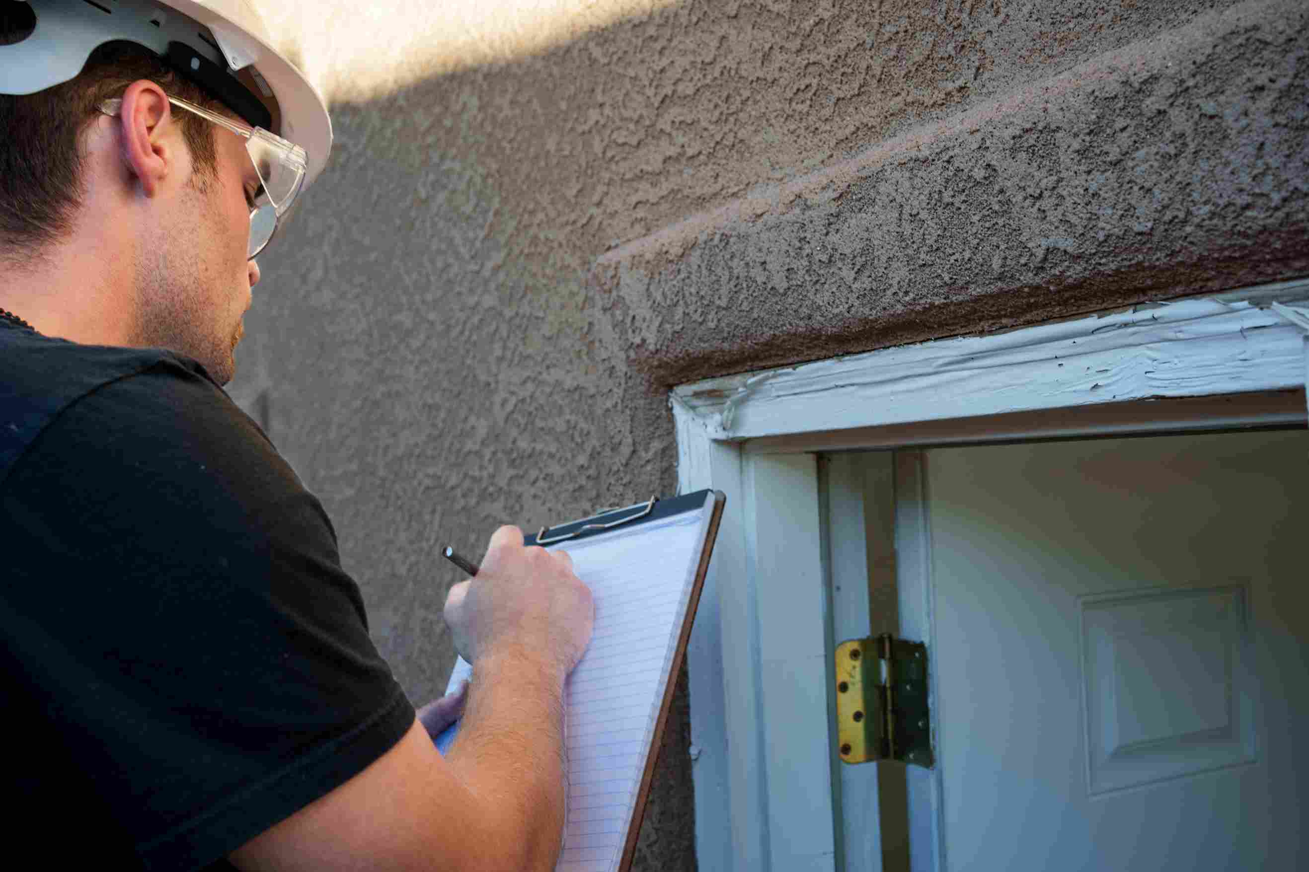 A person wearing a hard hat and safety glasses writes on a clipboard while inspecting a small exterior door.