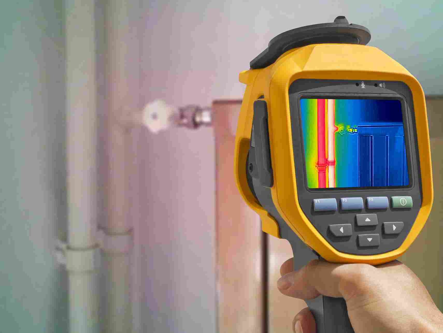 A person uses a handheld thermal imaging camera to detect temperature variations on a wall and pipes.