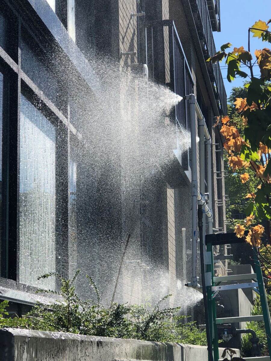 Water sprays from a pipe on the side of a building, with sunlight creating a mist effect. A ladder is propped against the structure, and trees are visible in the background.